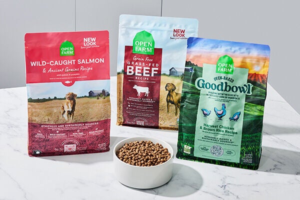 Three bags of open farm dry dog food, one wild caught salmon pink bag, one blue and red beef bag, and one green goodbowl bag behind a white bowl filled with kibble